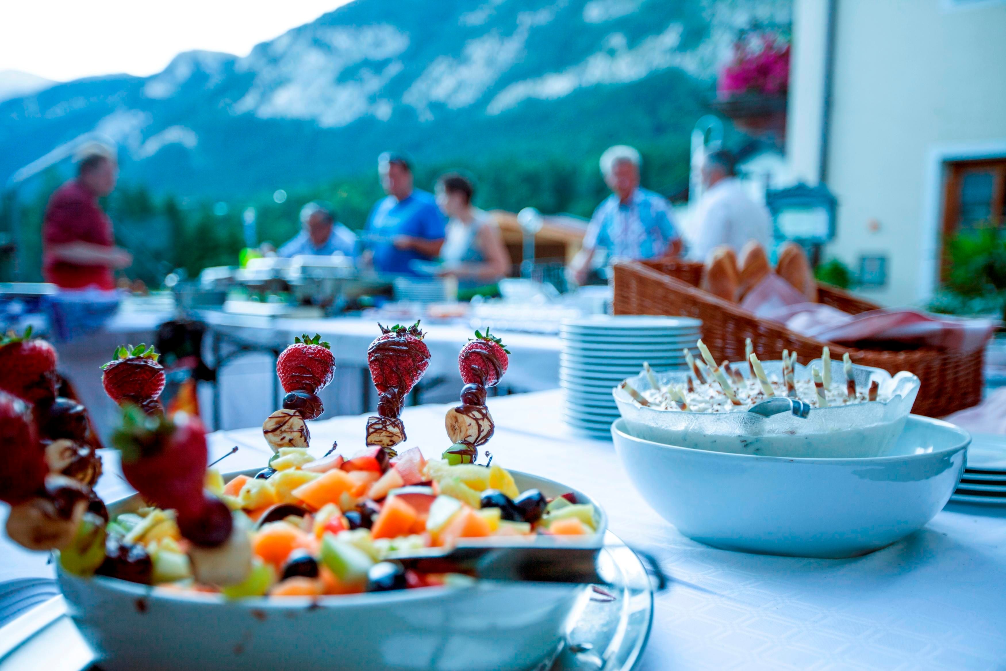 Ein festlich gedeckter Tisch mit bunten Obstspießen und einer Schüssel mit Snacks. Im Hintergrund sind Menschen und eine schöne Berglandschaft zu sehen.