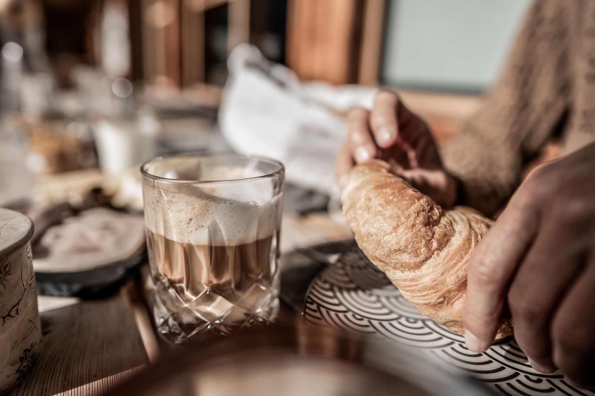 Ein Glas Kaffee steht auf einem Tisch. Eine Hand hält ein frisches Croissant.