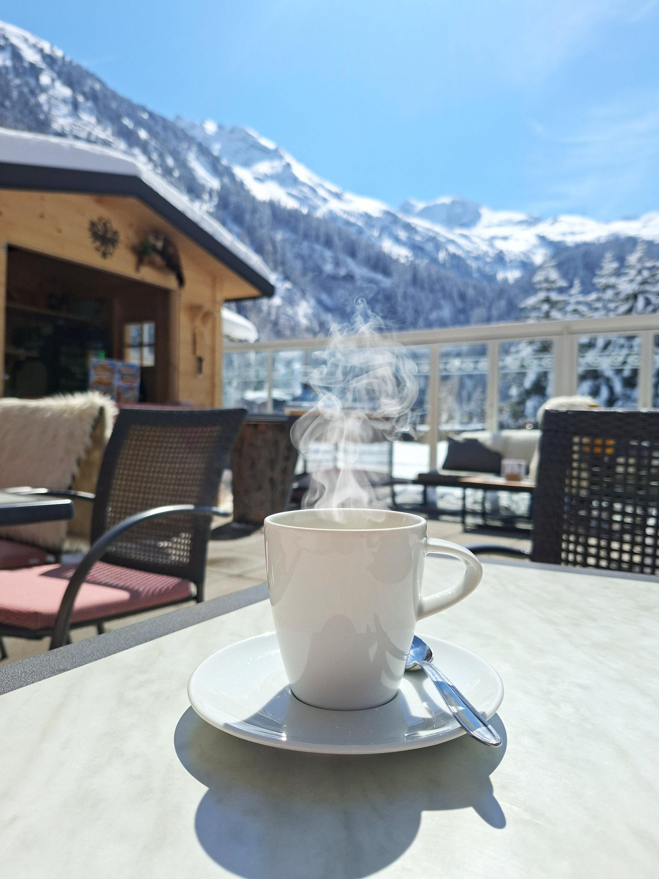 Eine dampfende Tasse Kaffee steht auf einem Tisch. Im Hintergrund sind schneebedeckte Berge und ein klarer blauer Himmel zu sehen.