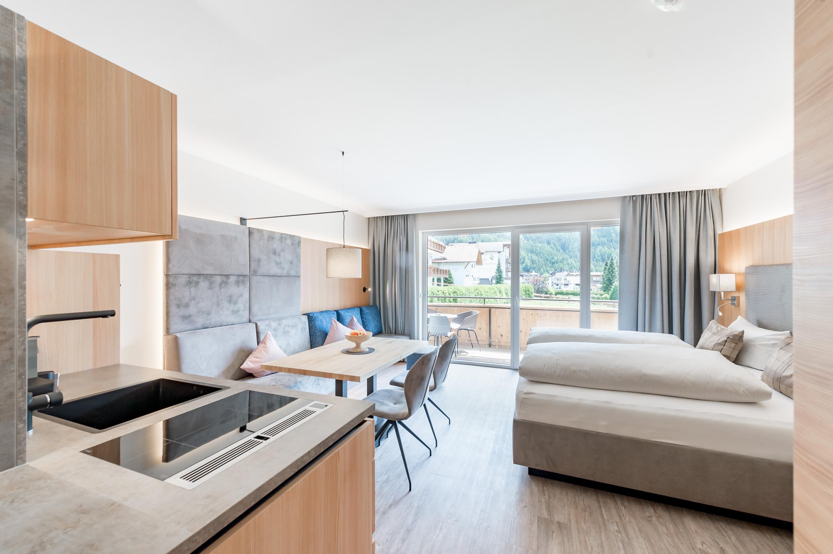 Ein modernes Zimmer mit einem großen Bett, einem Tisch und einer Küchenecke. Große Fenster bieten Blick auf die Natur.