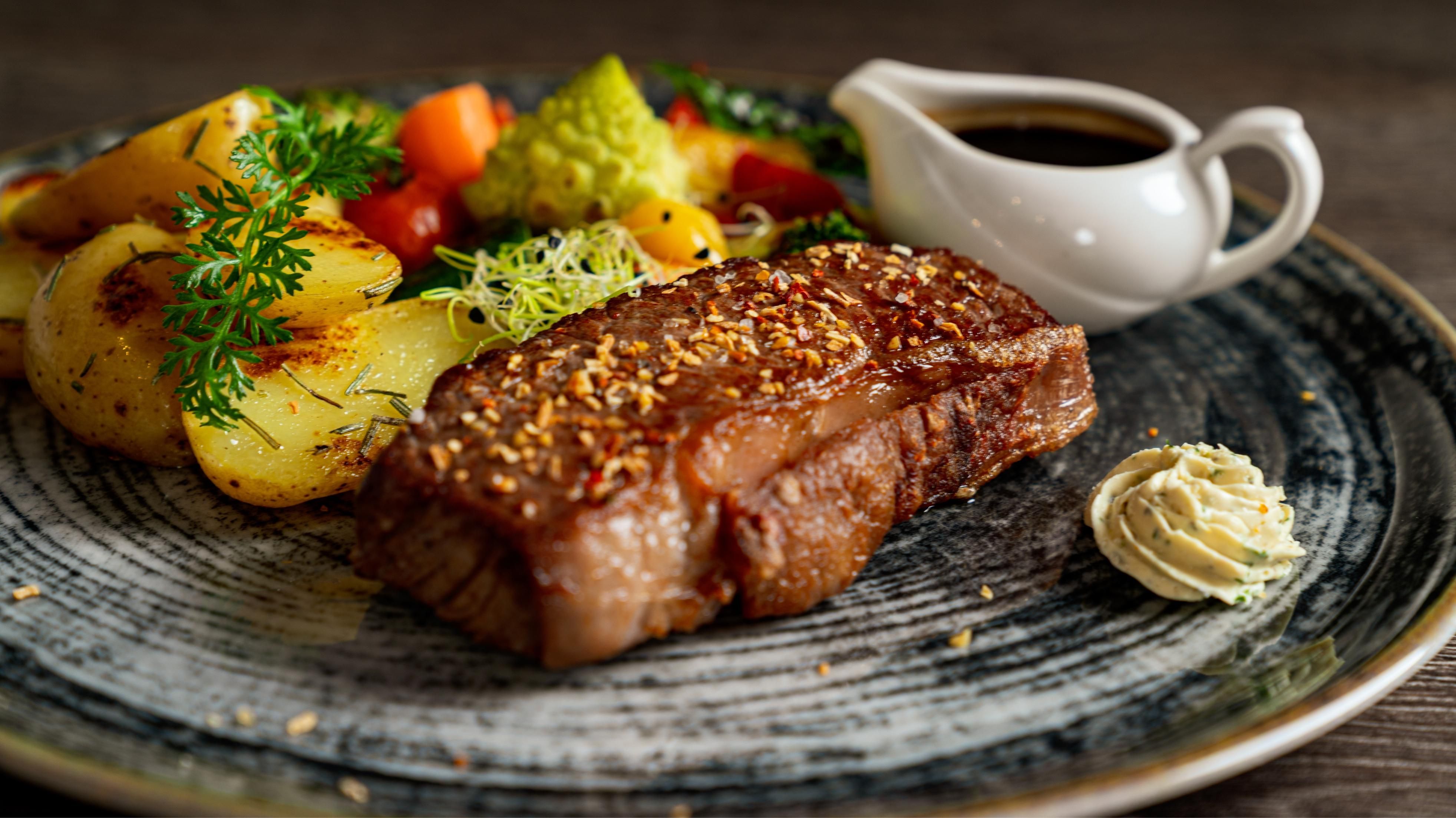 Ein köstliches Steak mit einer goldbraunen Kruste liegt auf einem Teller. Daneben befinden sich gebratene Kartoffeln und ein frischer Salat, begleitet von einer Sauce und etwas Kräuterbutter.