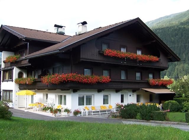 Ein gemütliches Holzhaus mit blühenden Balkonkästen. Vor dem Haus stehen gelbe Gartenmöbel und die Umgebung ist grün.