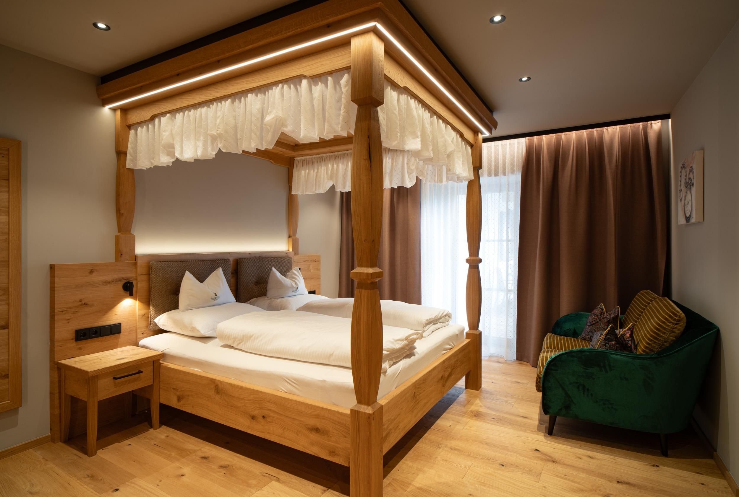 Ein gemütliches Schlafzimmer mit einem Himmelbett aus Holz. Es gibt einen kleinen Tisch und eine grüne Couch, sowie große Fenster mit Vorhängen.