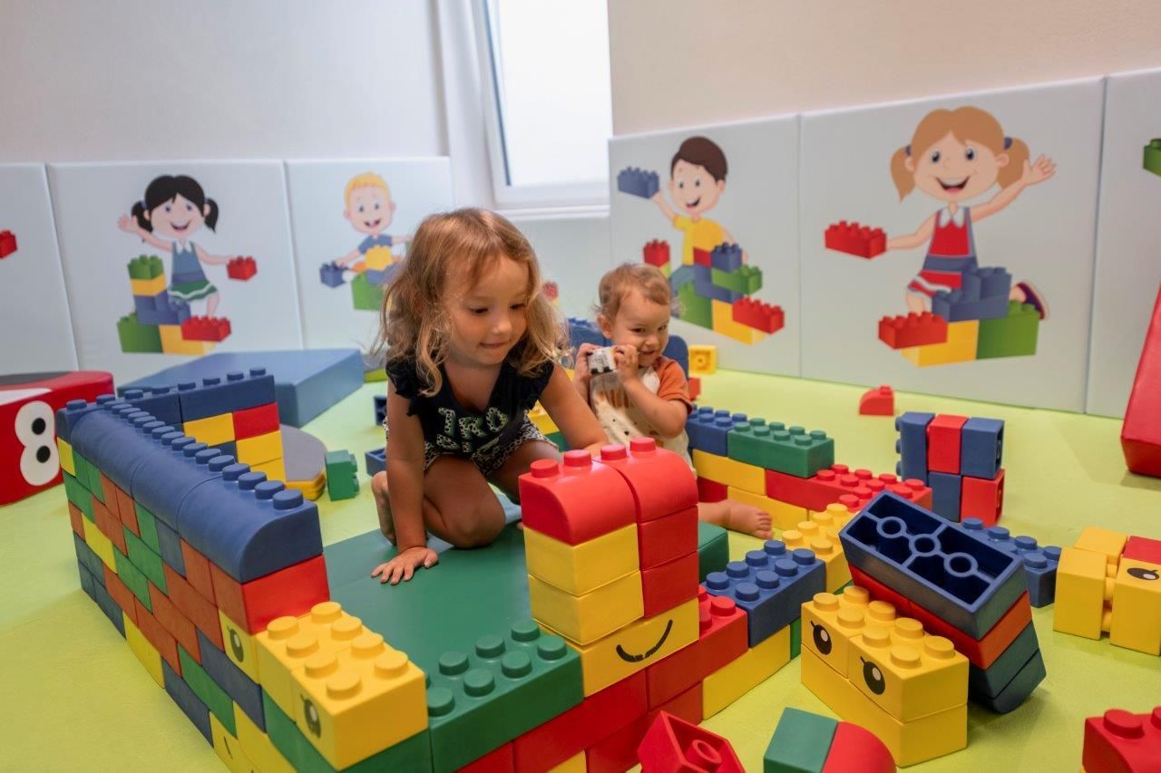 Zwei kleine Kinder spielen mit bunten Bausteinen in einem Spielezimmer. Im Hintergrund sind fröhliche Wandbilder von anderen Kindern zu sehen.