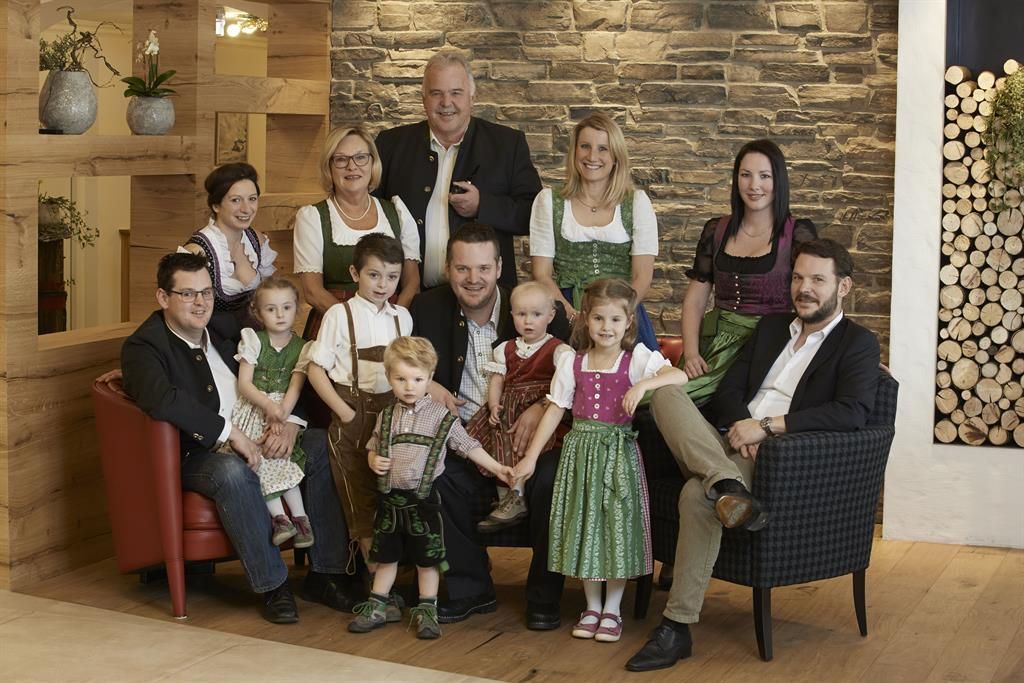 Eine Gruppe von Menschen in traditioneller Tracht steht und sitzt in einem gemütlichen Raum. Im Hintergrund sind Holzdekorationen und eine natürliche Umgebung sichtbar.