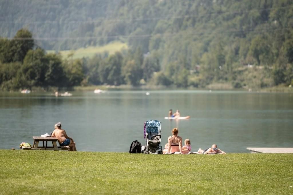 Ein ruhiger See mit grünem Rasen und einigen Menschen, die sich entspannen. Im Hintergrund sind Paddler auf dem Wasser zu sehen.