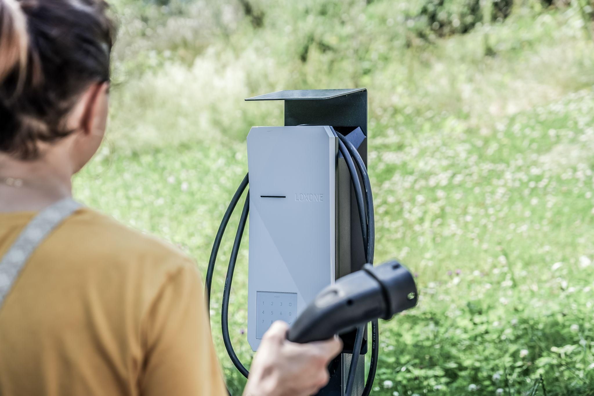 Eine Person steht vor einer Ladestation für Elektrofahrzeuge. Im Hintergrund sind grüne Wiesen und Bäume zu sehen.