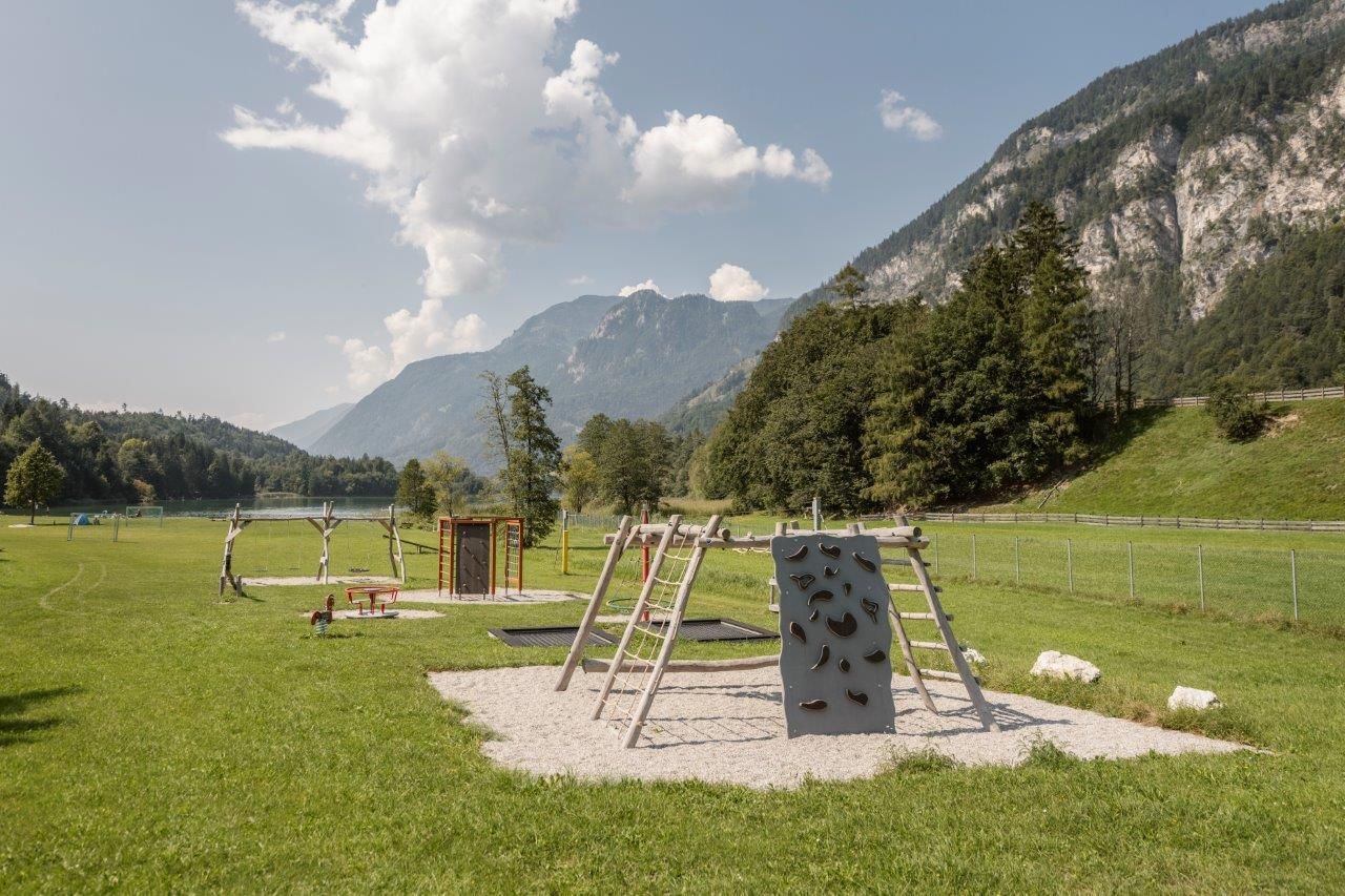 Ein Spielplatz in einer grünen Wiese mit Bergen im Hintergrund. Es gibt Klettergerüste und einen Kletterfelsen zum Spielen.