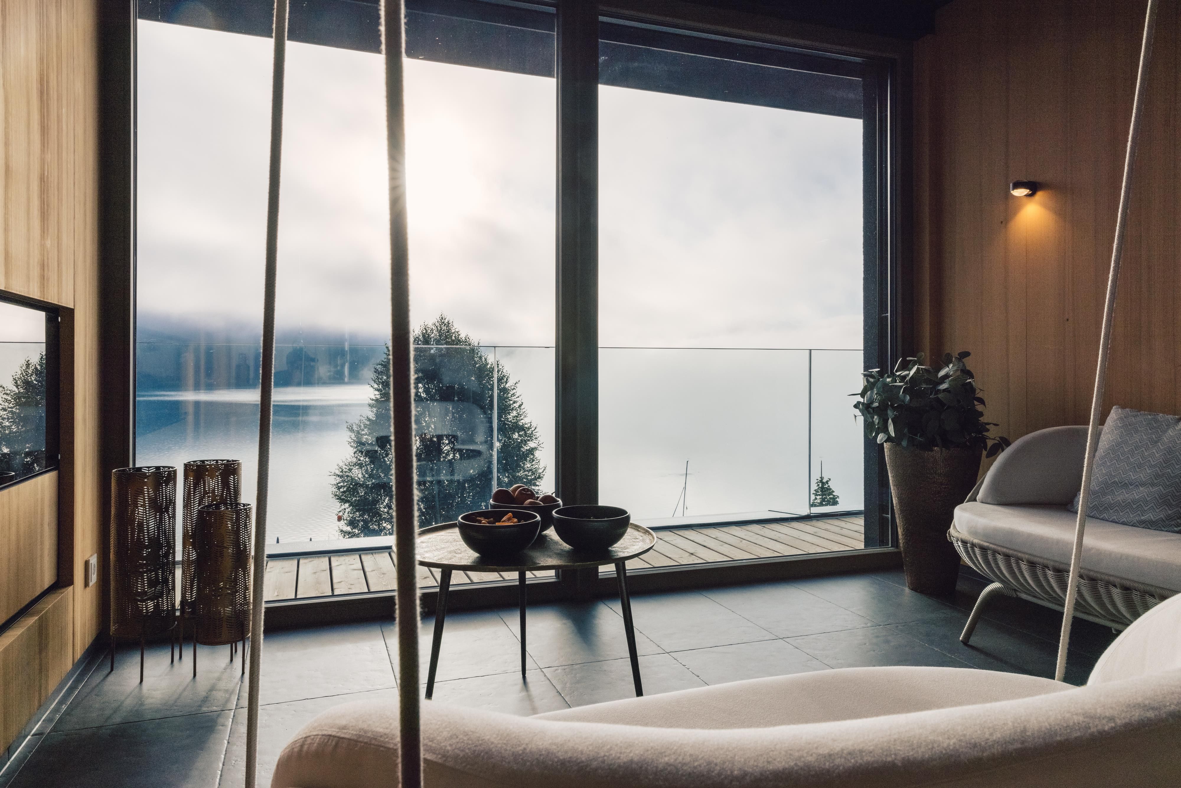 Ein stilvolles Wohnzimmer mit großen Fenstern und Blick auf einen ruhigen See. Die Holzwände und moderne Möbel schaffen eine gemütliche Atmosphäre.