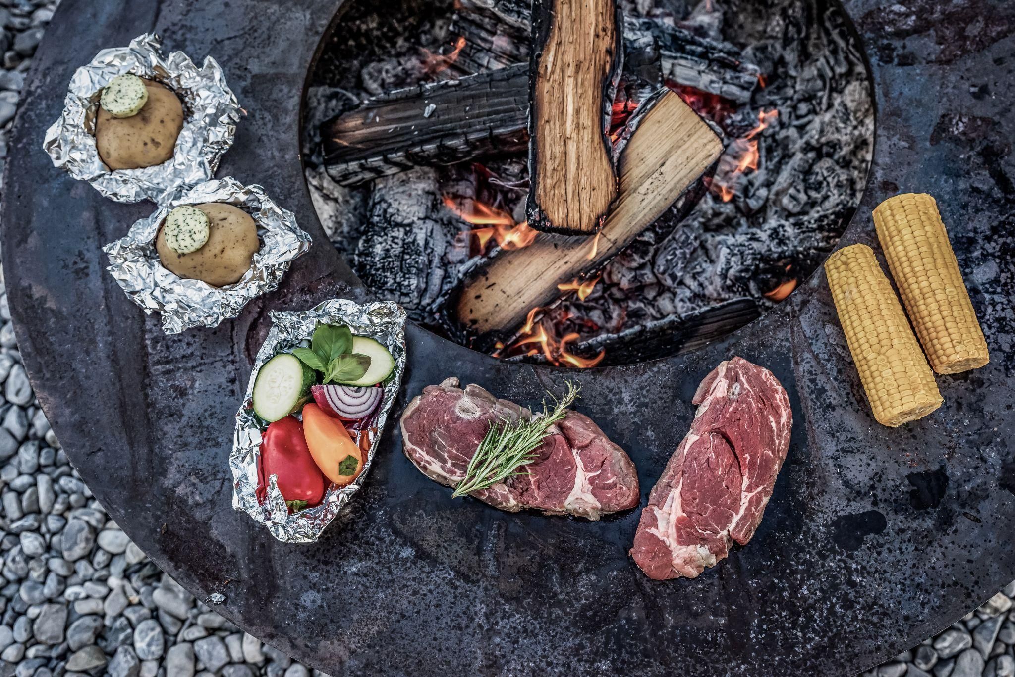 Auf dem Bild sind mehrere Lebensmittel zum Grillen zu sehen, darunter frisches Gemüse, Fleischstücke und Kartoffeln, die in Folie gewickelt sind. Im Hintergrund brennen Holzscheite über offenem Feuer.