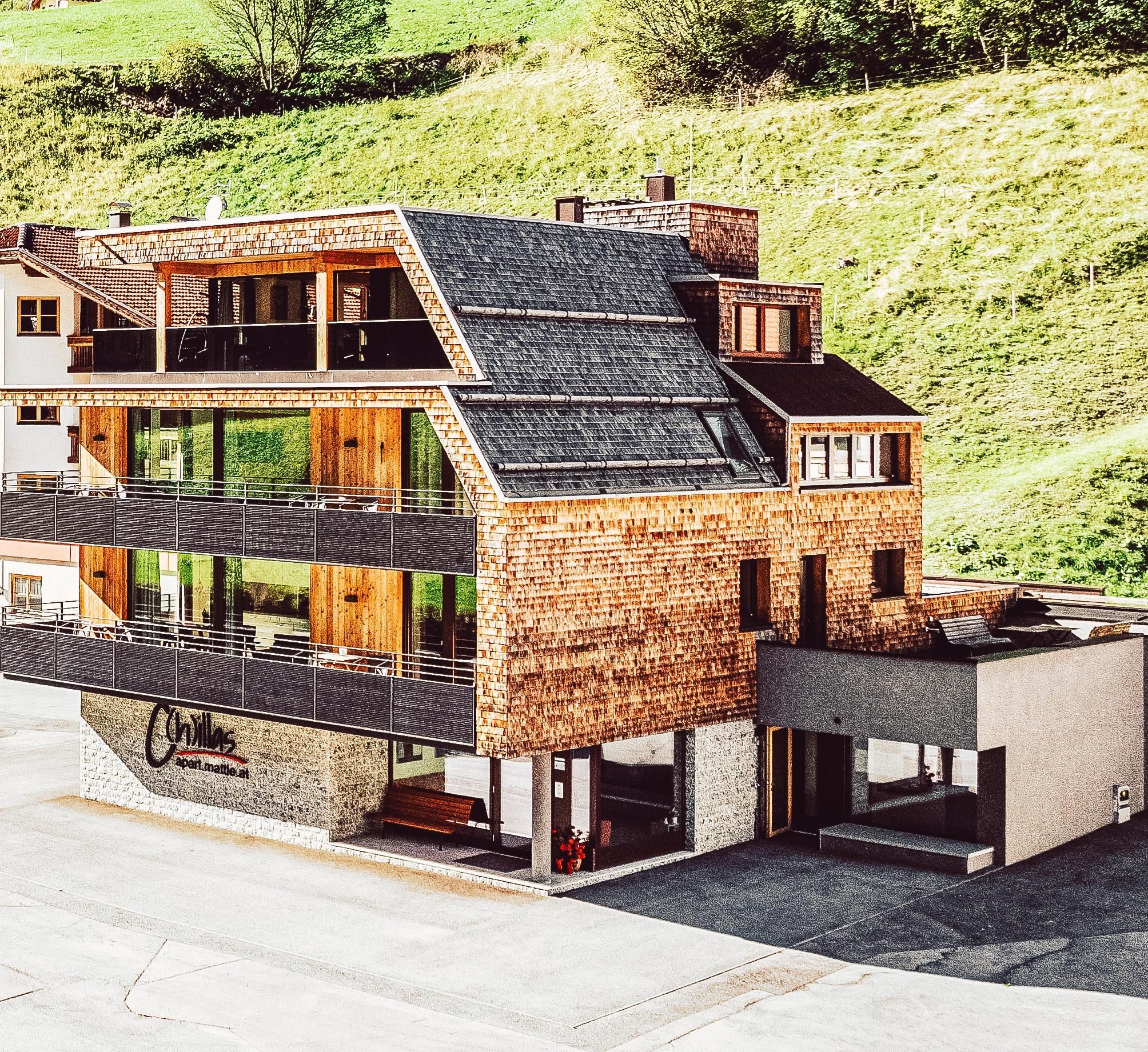 Ein modernes Gebäude mit Holz- und Steinfassade inmitten einer grünen Landschaft. Es verfügt über große Fenster und Balkone, die eine helle und einladende Atmosphäre schaffen.