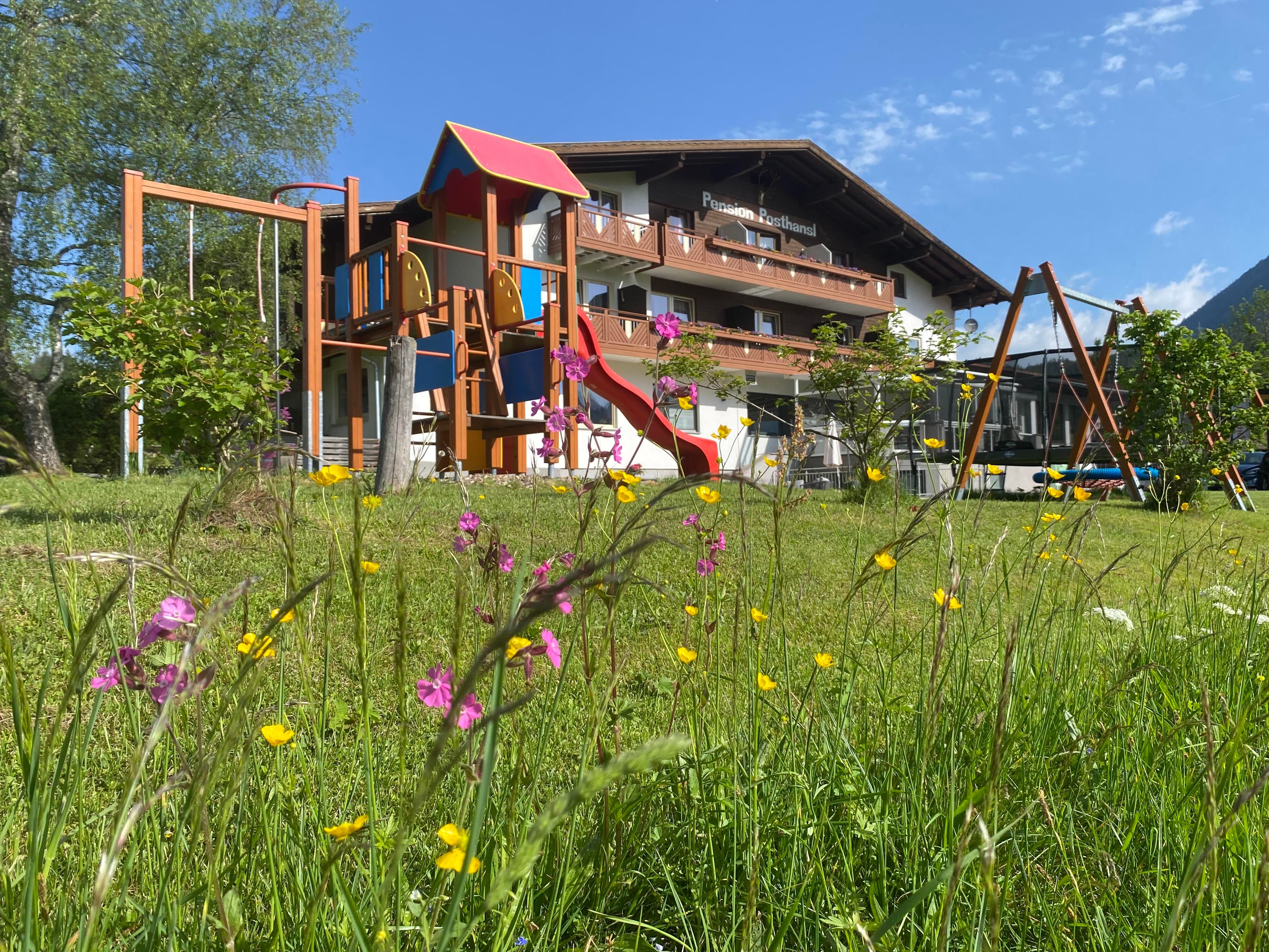 Ein Garten mit bunten Blumen und einem Spielplatz mit Rutsche und Schaukeln. Im Hintergrund steht ein gemütliches Haus unter blauem Himmel.