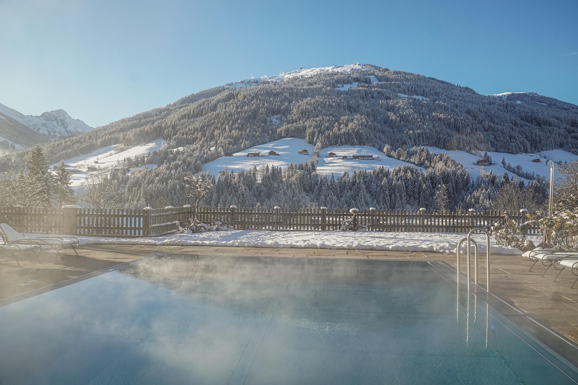 Ein beheizter Pool mit dampfendem Wasser vor einer schneebedeckten Berglandschaft. Der Himmel ist klar und die Aussicht ist atemberaubend.