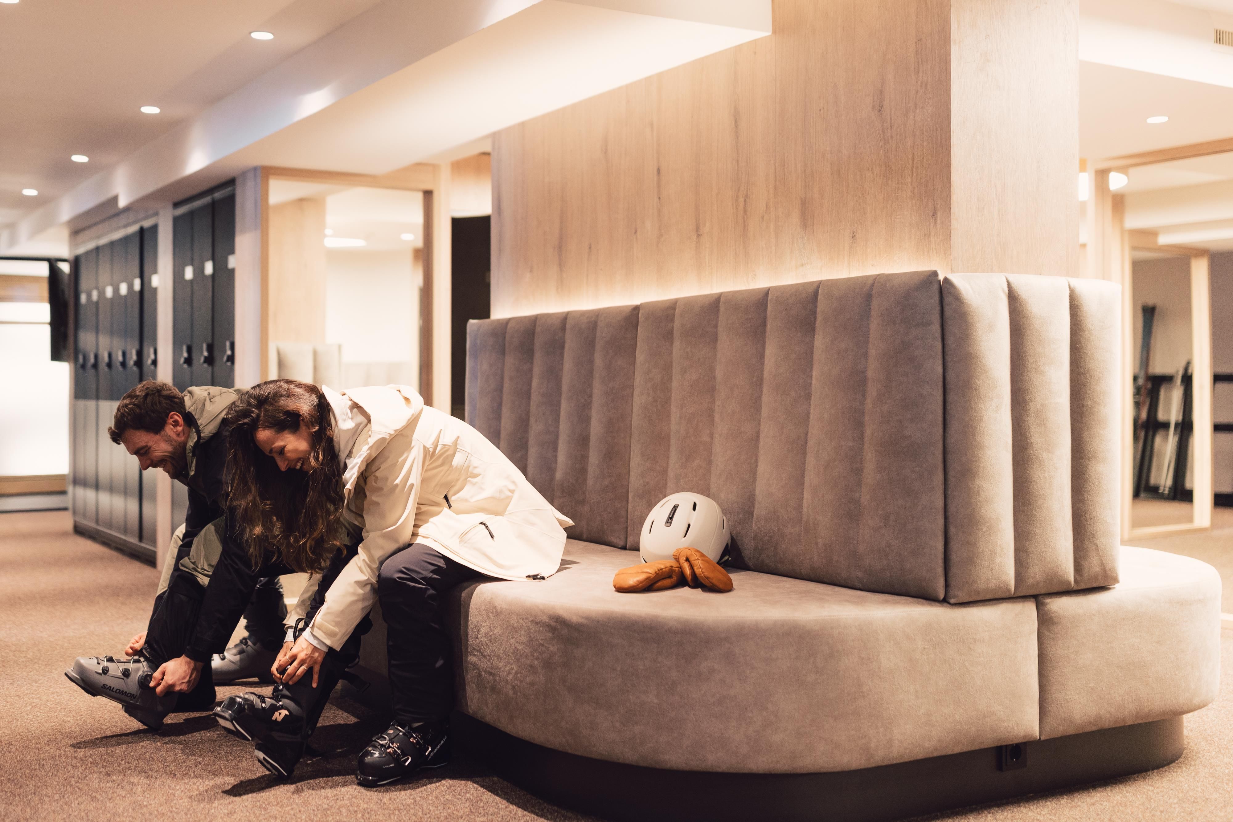 Zwei Personen schnüren sich ihre Skischuhe in einem modernen Umkleideraum. Im Hintergrund sind Schließfächer zu sehen.