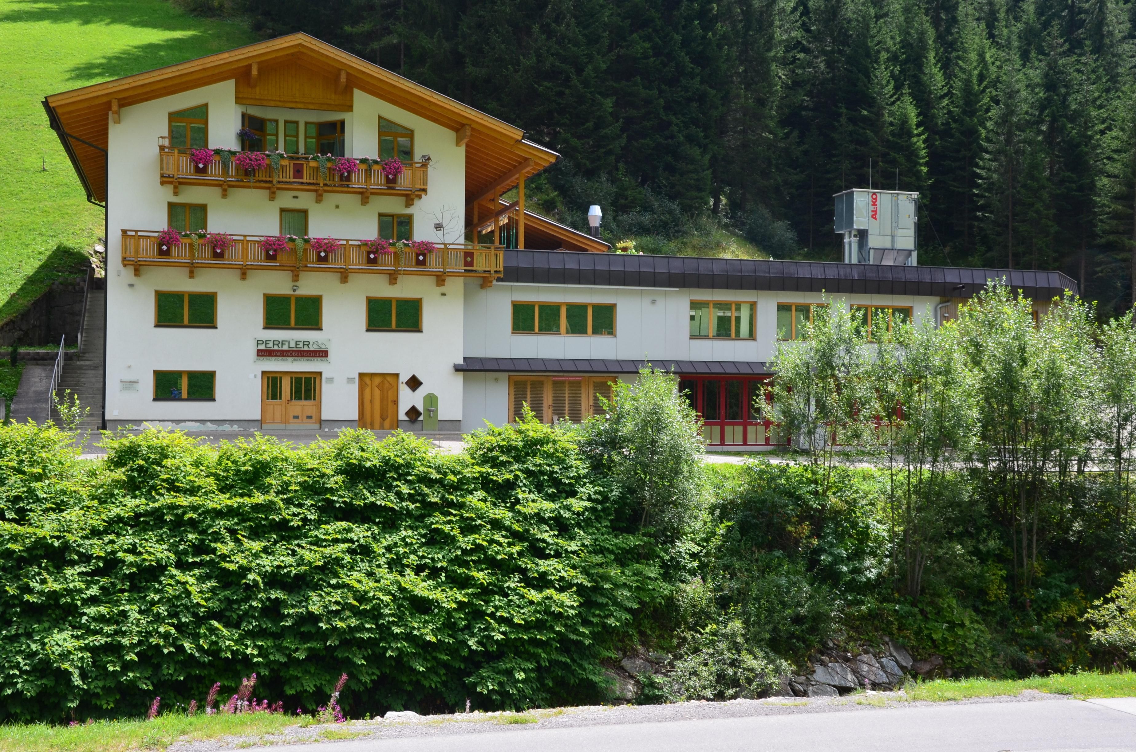Ein großes Gebäude im alpinen Stil, umgeben von Bäumen und grünen Wiesen. Die Ferienwohnung befindet sich im 1. Stockwerk mit Balkon und Zugang zur Terrasse.