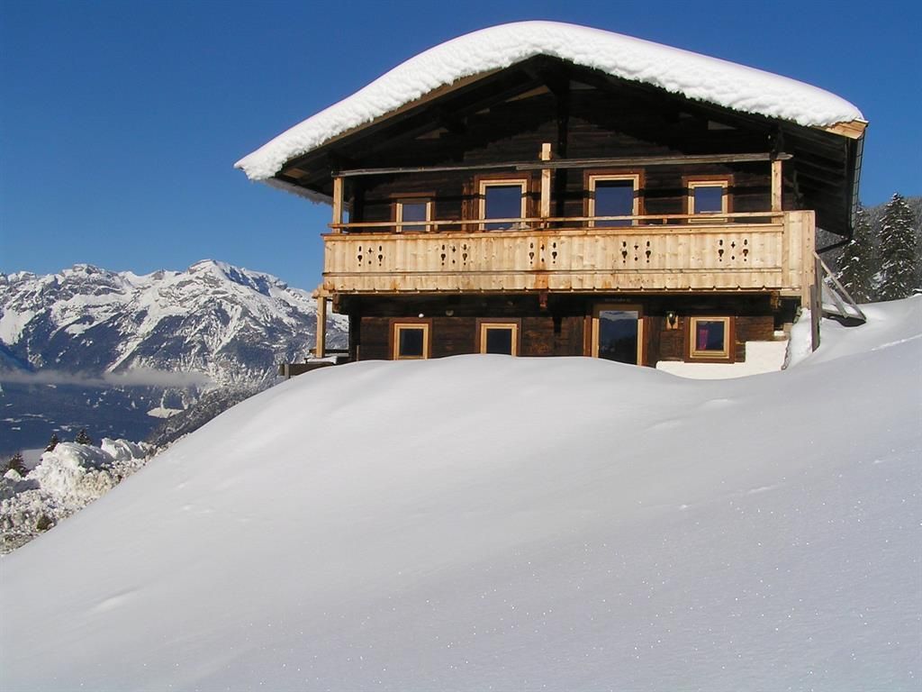 Ein gemütliches Holzhaus im Schnee mit einem großen Balkon. Im Hintergrund sind majestätische Berge unter einem klaren blauen Himmel zu sehen.