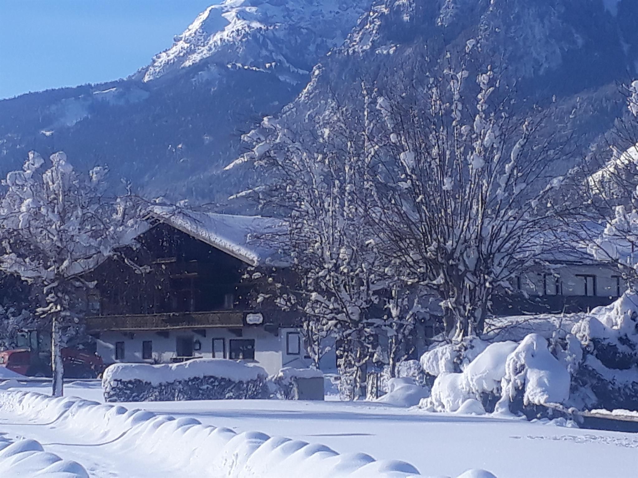 Eine verschneite Landschaft mit einem Chalet und schneebedeckten Bäumen. Im Hintergrund ragen majestätische Berge in den blauen Himmel.