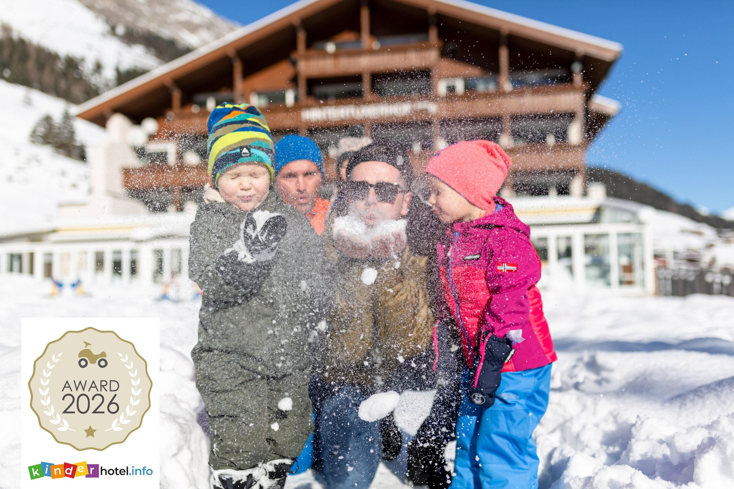 Eine Familie spielt im Schnee vor einem großen Holzgebäude. Sie lachen und werfen Schnee in die Luft, während die Sonne scheint.
