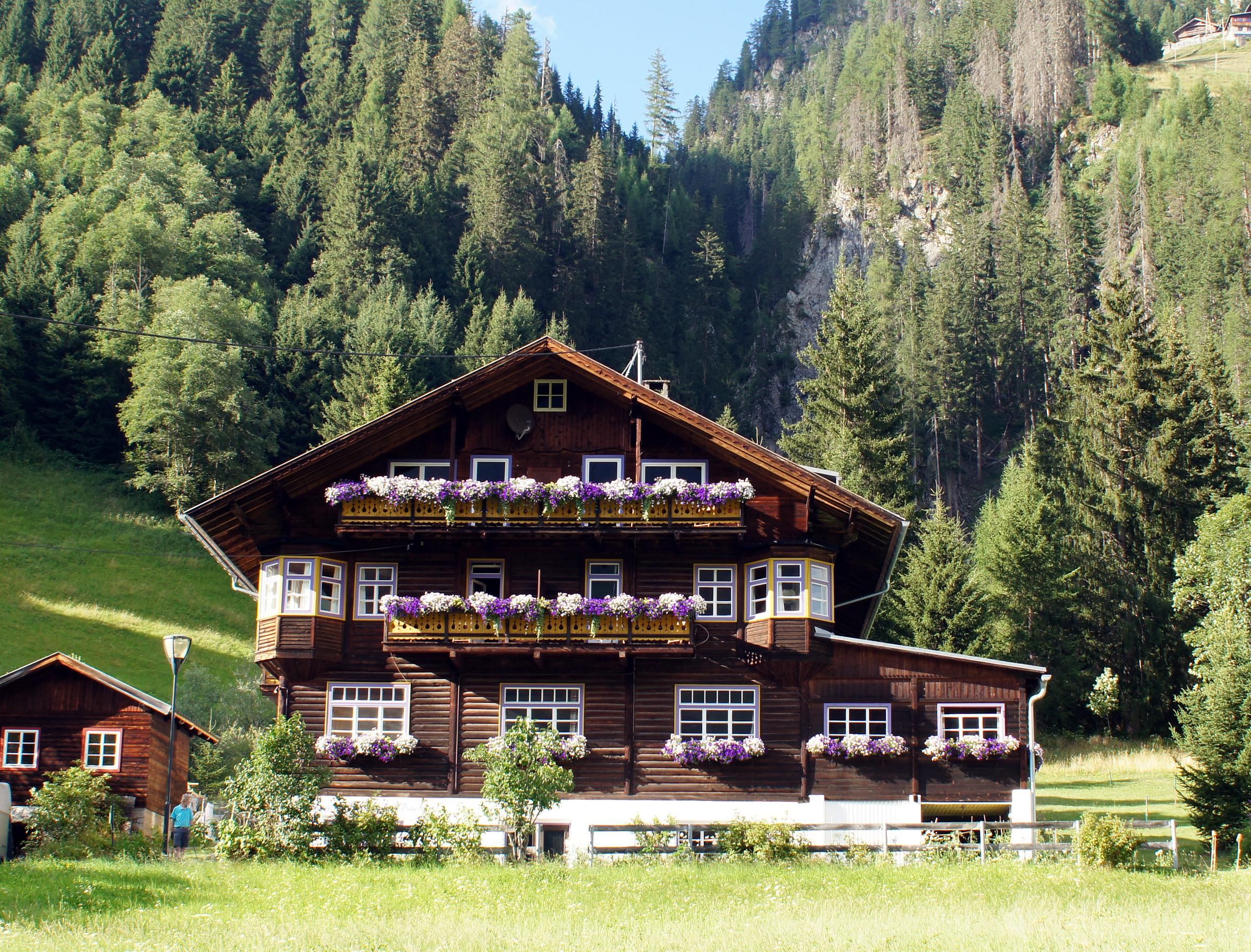 Das Refugium Tegischer Bachl von vorne im Sommer: auf einem gemauerten Kellergeschoß, das weiß gestrichen ist, steht ein Holzblockbau in dunkelbraun. Im Erdgeschoß sind große, weiß umrandete Kastenfenster, das erste und zweite Obergeschoß haben jeweils Balkone. An allen Fenstern und Balkonen sind Blumen. Links vom Haupthaus steht noch ein kleineres Häuschen. Vor dem Haus ist eine Wiese, dahinter steigt der Berg an und es stehen Bäume.