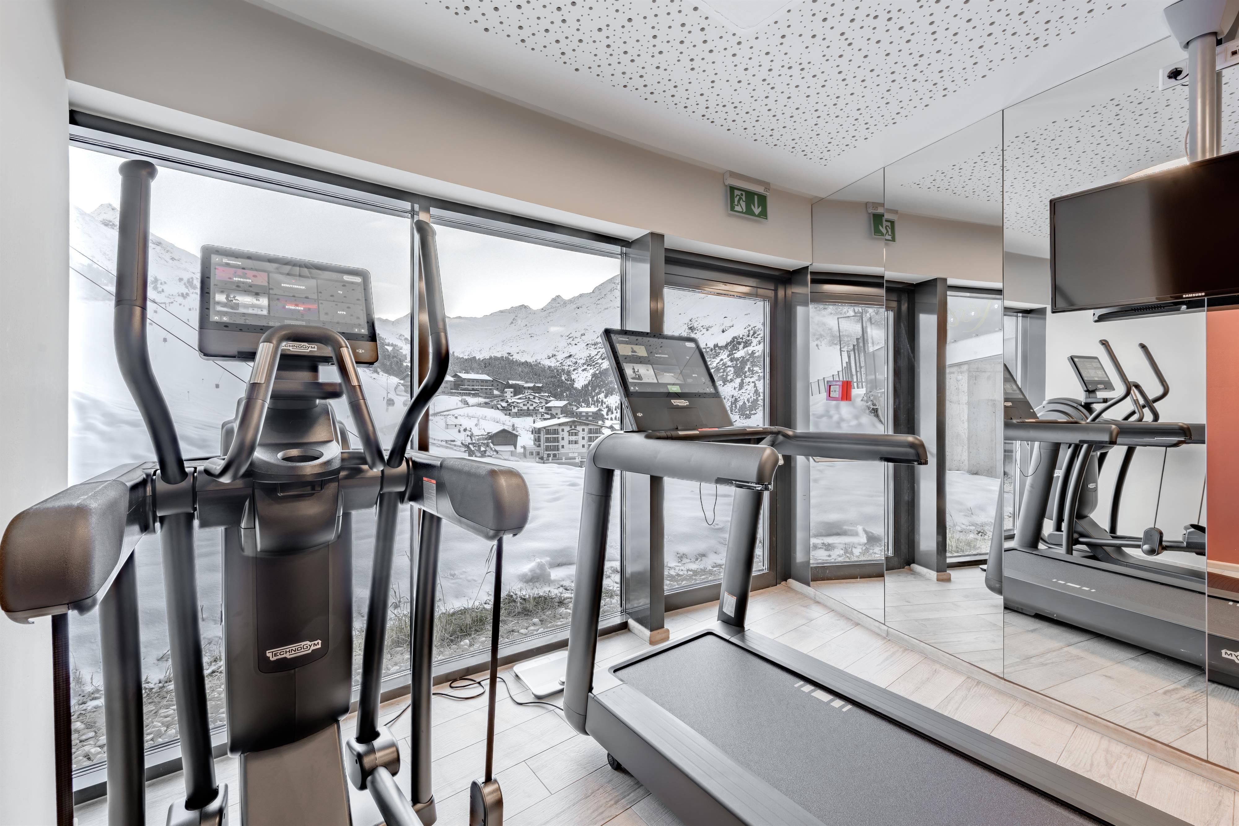 Ein moderner Fitnessraum mit Blick auf die Berge. Vor einem großen Fenster stehen ein Laufband und ein Crosstrainer.