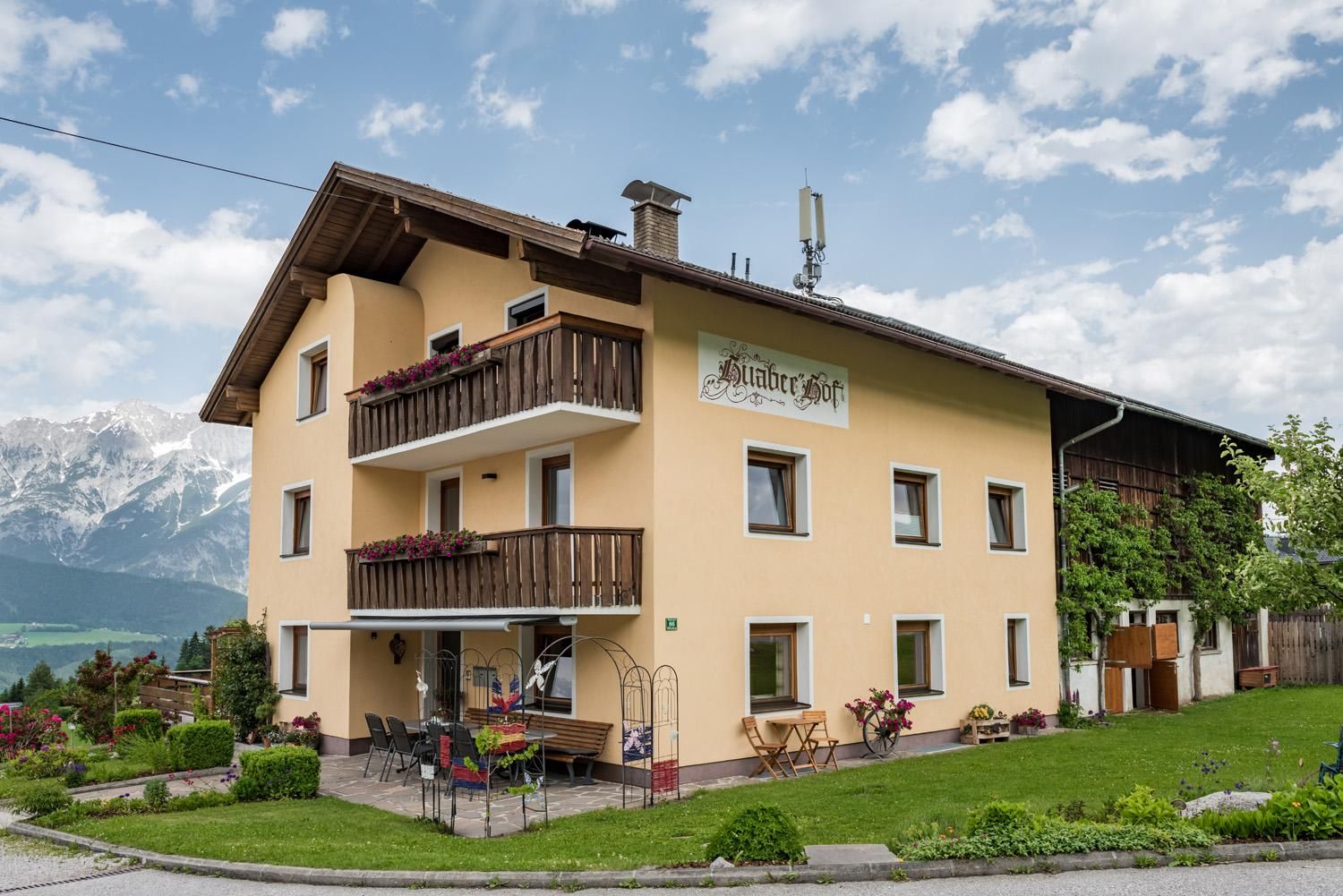 Ein modernes Wohnhaus mit einem Balkon und Blumen. Im Hintergrund sind Berge und ein blauer Himmel zu sehen.