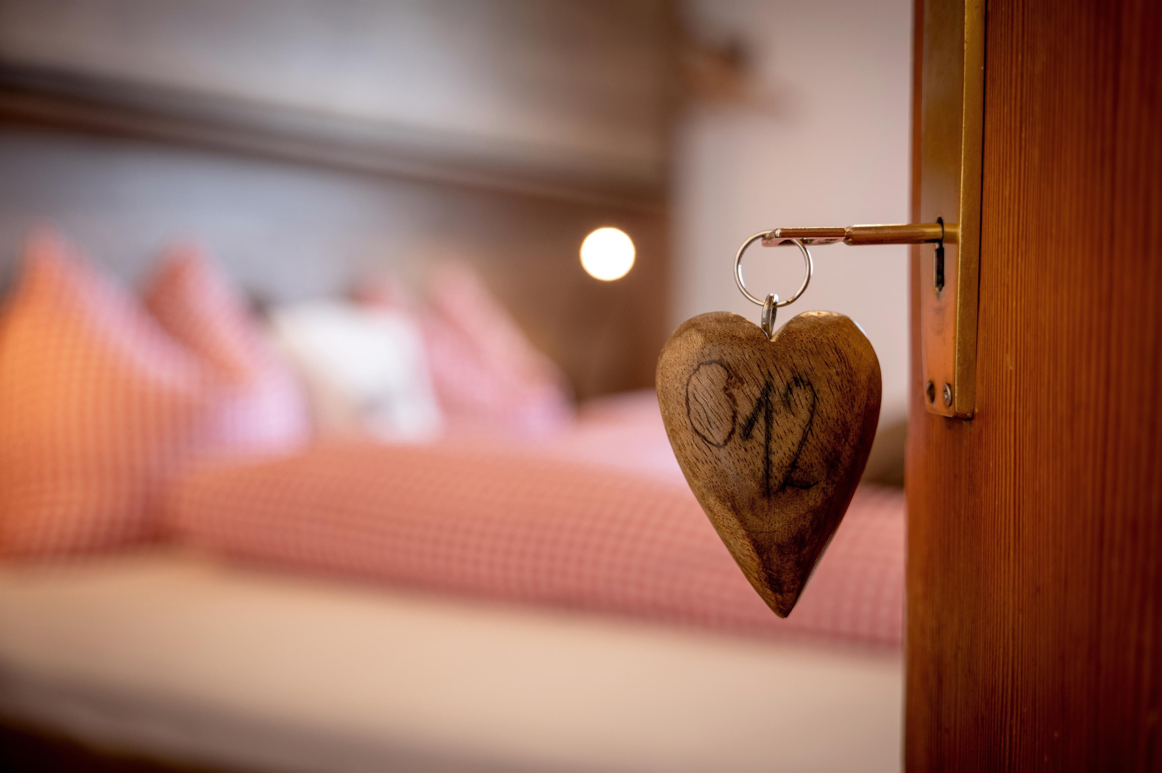 Ein Holzherz mit der Zahl "02" hängt an einer Zimmertür. Im Hintergrund ist ein gemütliches Schlafzimmer mit karierten Kissen zu sehen.