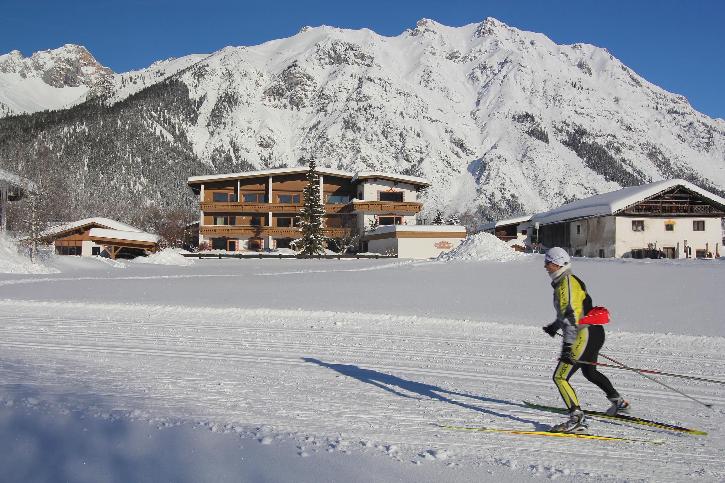 Eine Person fährt Ski auf einer schneebedeckten Fläche mit einem malerischen Berg im Hintergrund. Im Hintergrund sind außerdem einige Gebäude zu sehen.