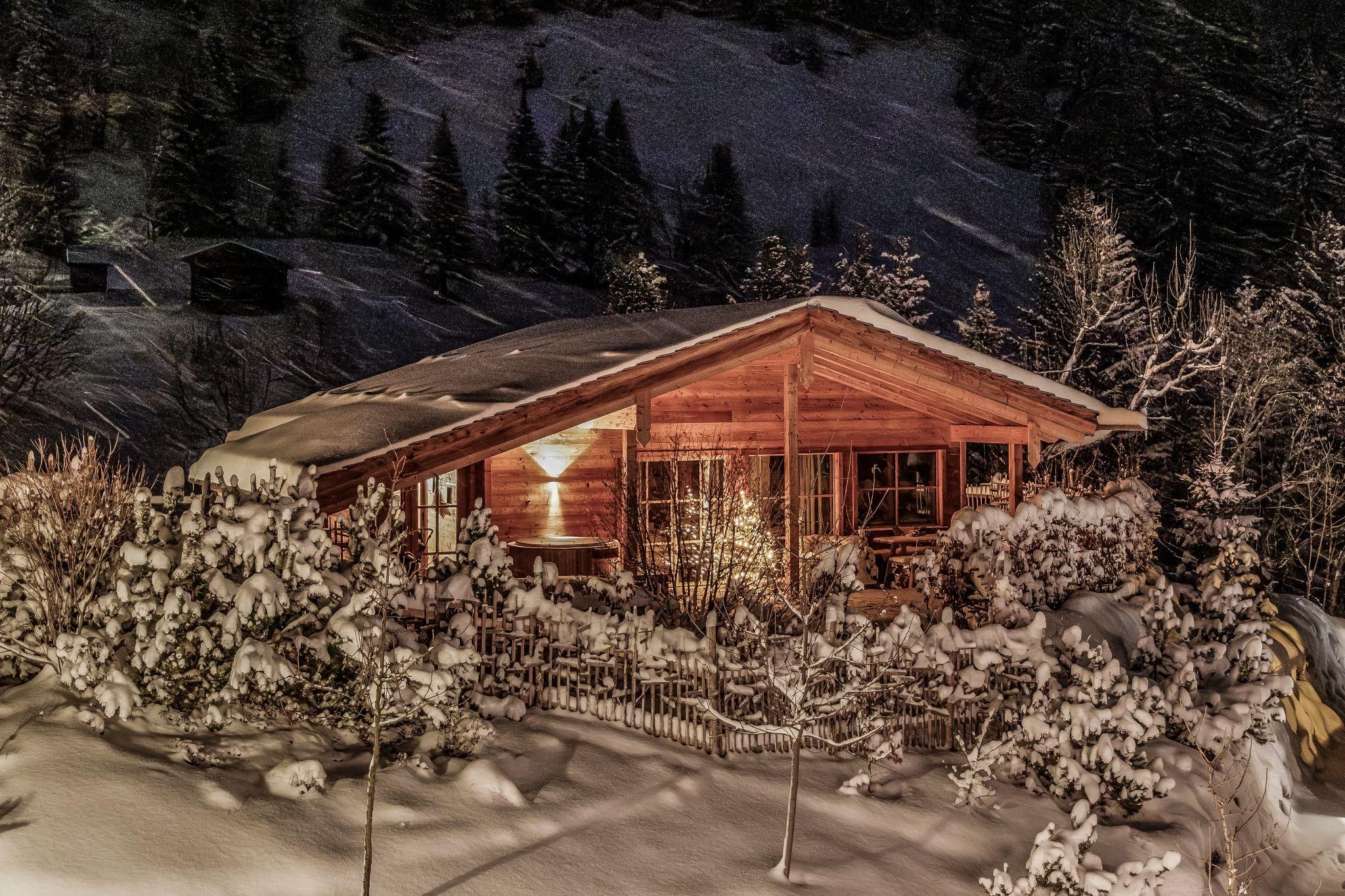 Ein gemütliches Chalet in einer verschneiten Landschaft bei Nacht. Sanfte Lichter strahlen durch die Fenster und schaffen eine warme Atmosphäre.