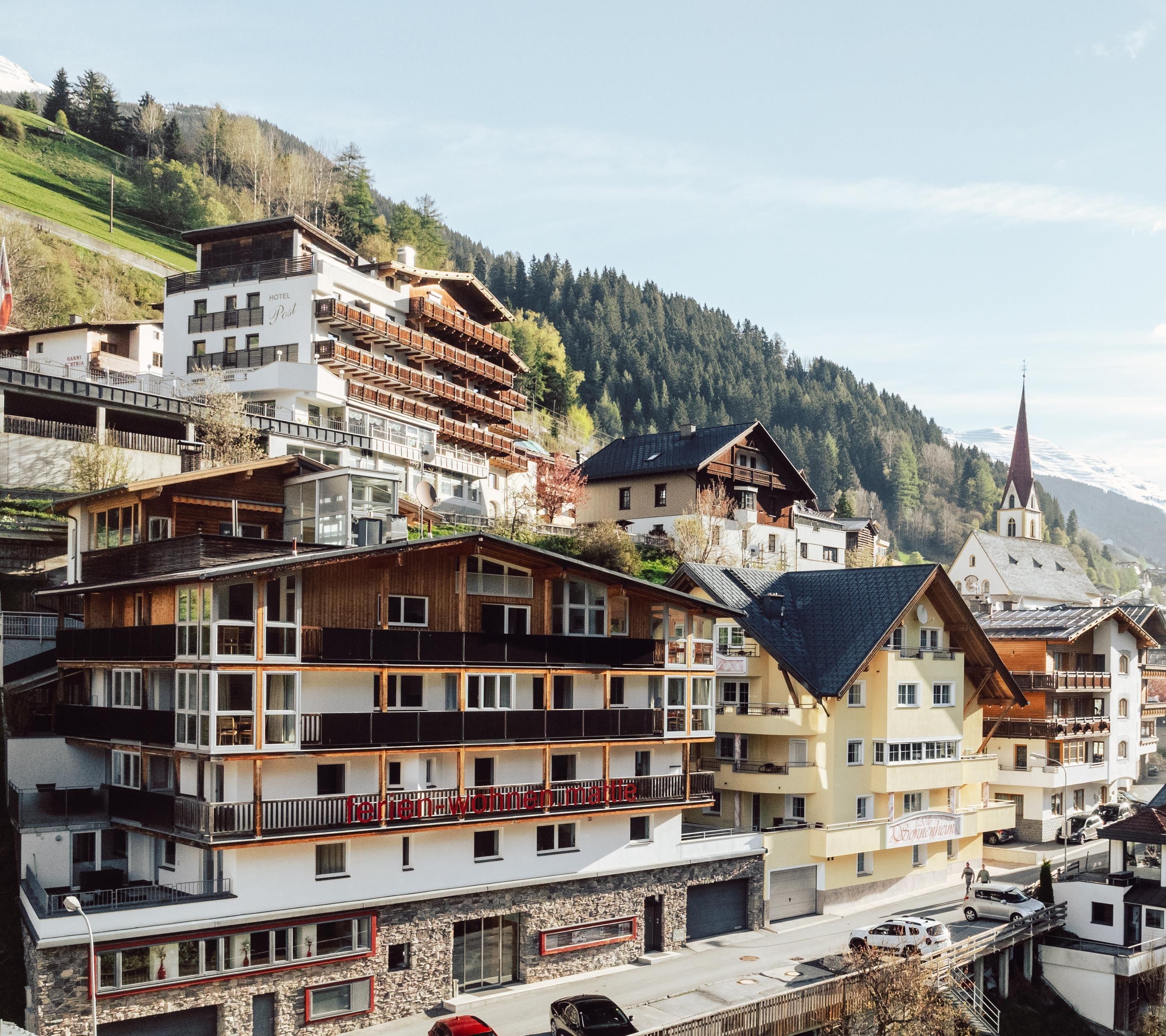 Eine malerische Berglandschaft mit mehreren modernen Chalets und traditionellen Gebäuden. Die Häuser liegen an einem Hang, umgeben von Wäldern und Bergen.