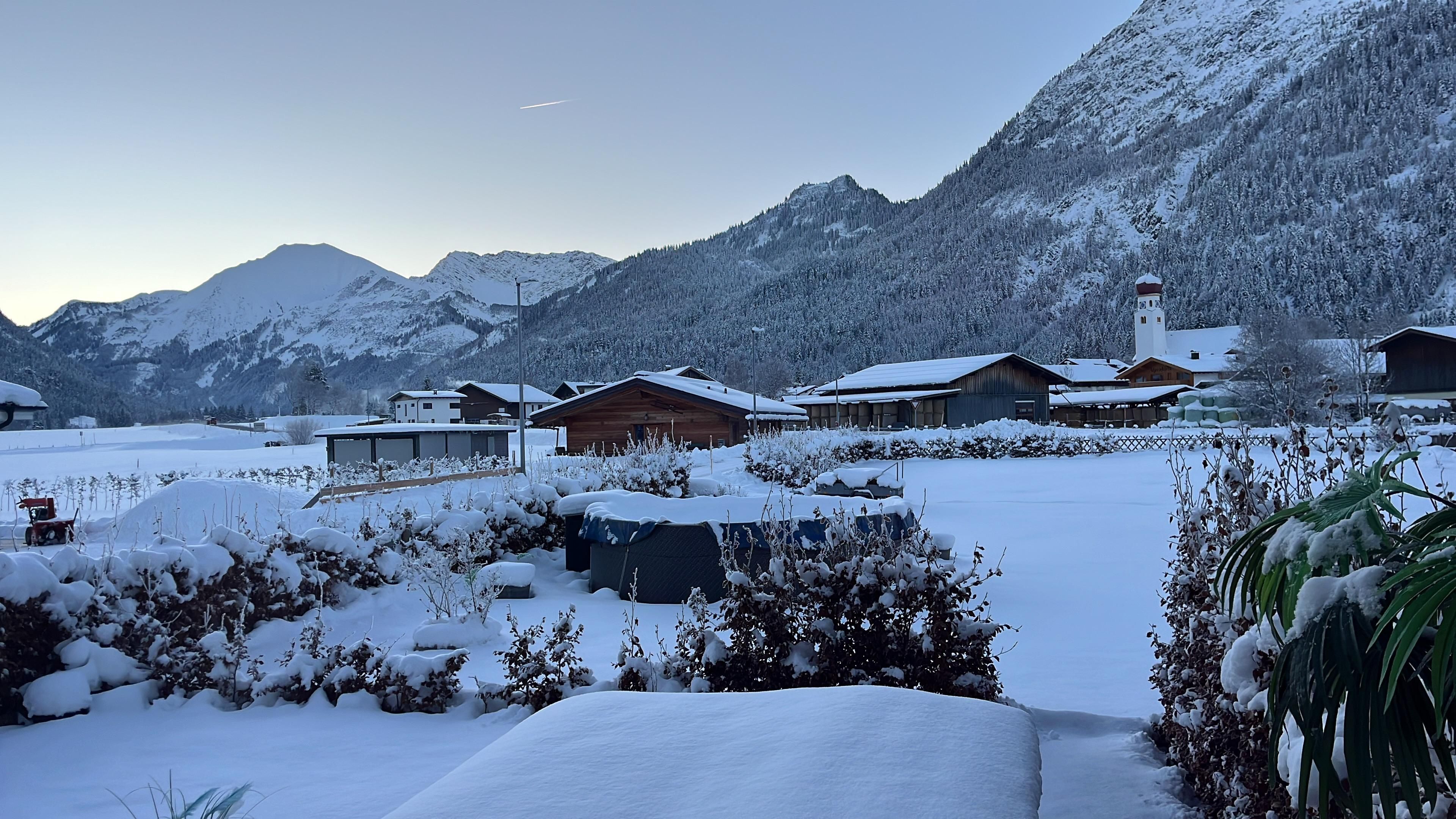 Eine verschneite Landschaft mit Bergen im Hintergrund und traditionellen Holzhäusern. Der Himmel ist klar und es liegt viel Schnee auf dem Boden.
