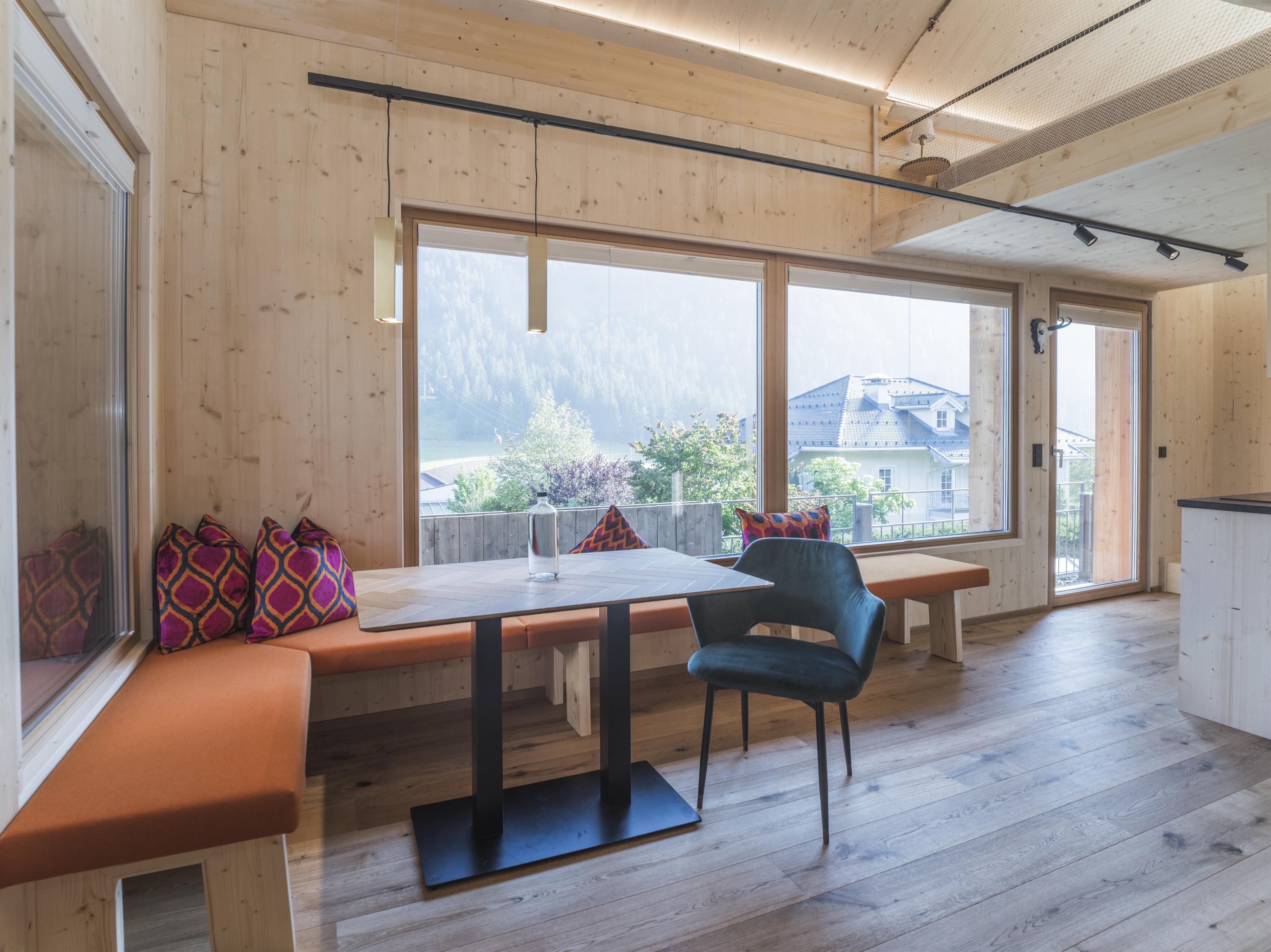 Ein modernes Raum mit Holzverkleidung und großen Fenstern. Ein Esstisch mit Stühlen und eine gemütliche Bank mit Kissen sorgen für eine einladende Atmosphäre.