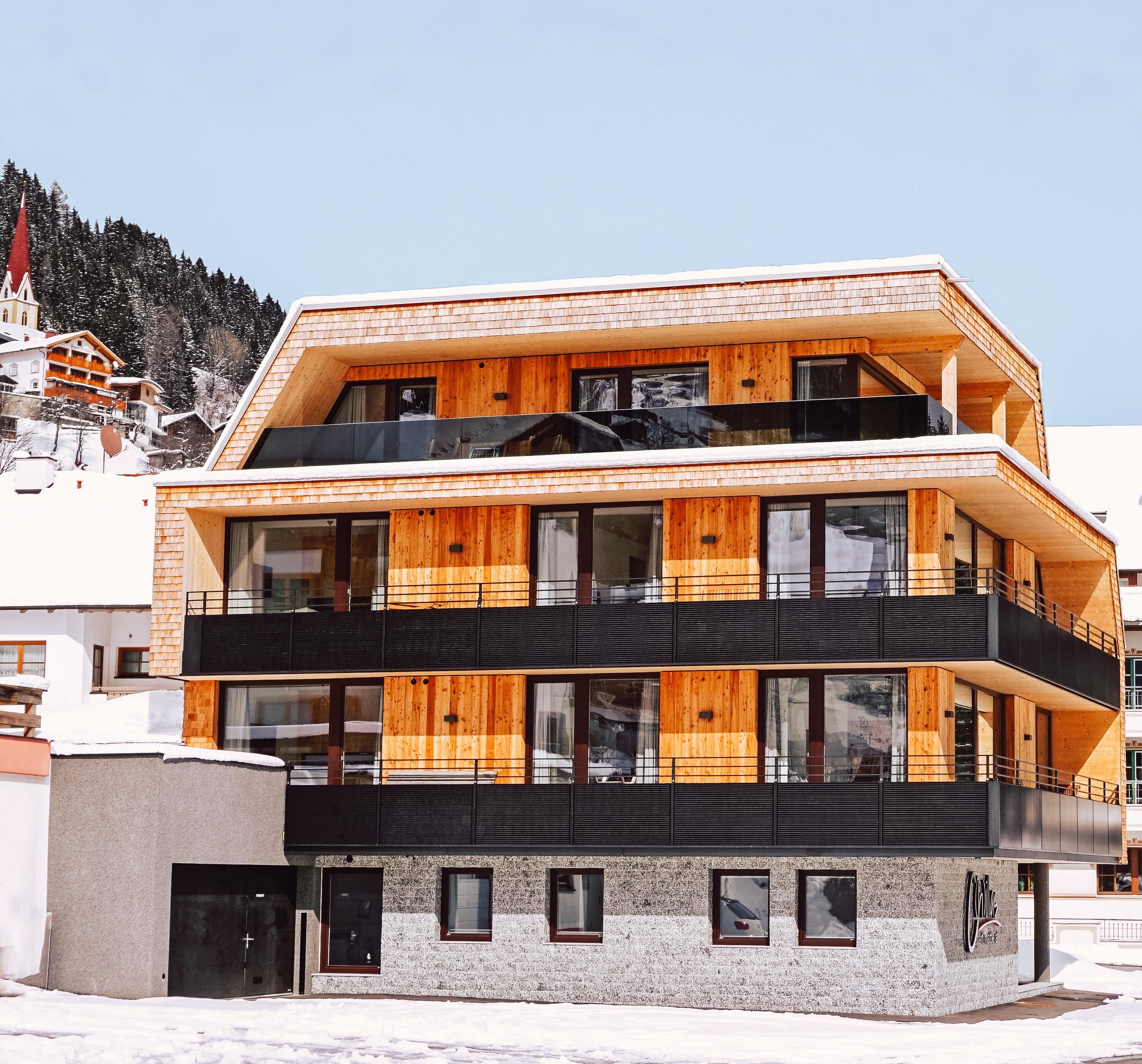 Ein modernes Gebäude mit Holzverkleidung in einer winterlichen Umgebung. Schnee liegt auf dem Boden und der Blick reicht auf die bewaldeten Berge im Hintergrund.