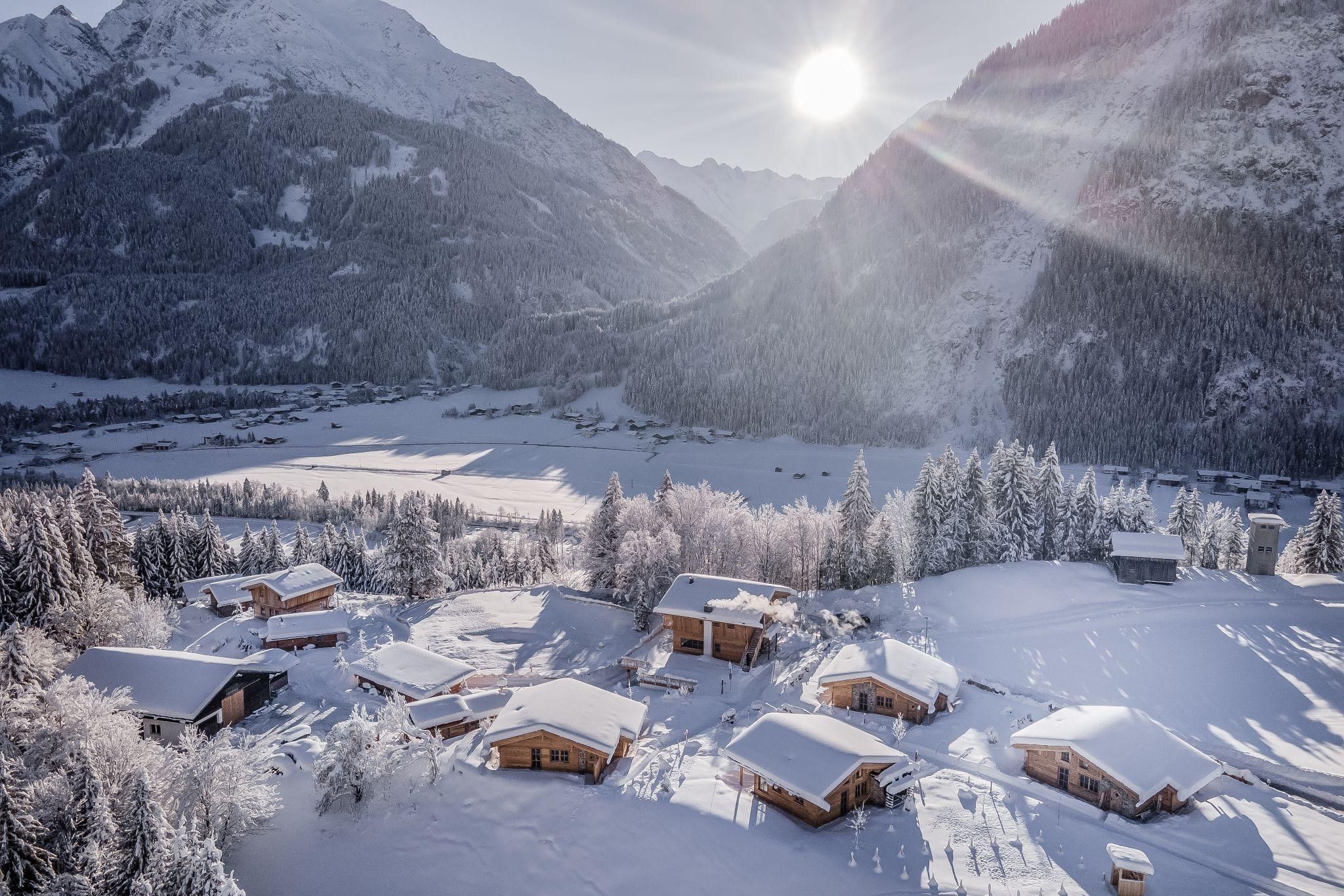 Eine malerische Winterlandschaft mit schneebedeckten Chalets und hohen Bergen. Die Sonne scheint hell über das ruhige Tal.