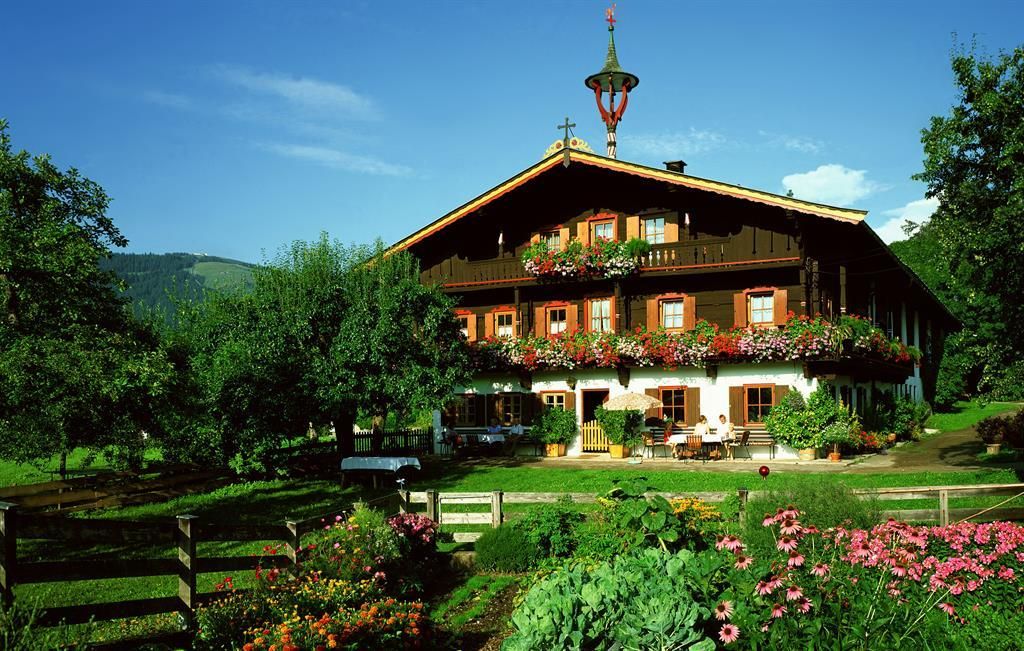 Ein traditionelles, bayerisches Haus mit einem schönen Balkon voller Blumen. Die Umgebung ist grün und idyllisch, umgeben von Bäumen und blühenden Gartenpflanzen.