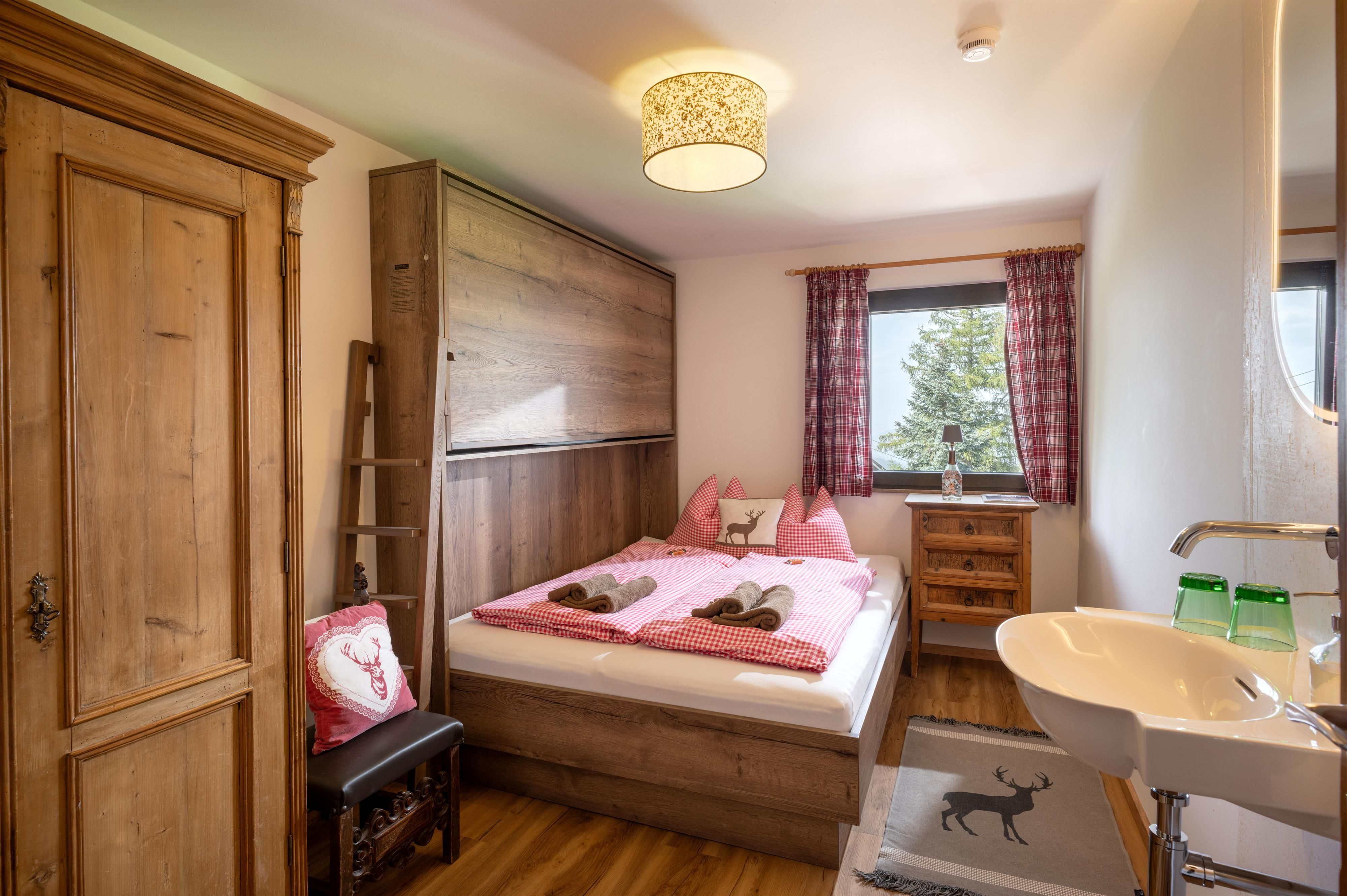 Ein gemütliches Schlafzimmer mit einem Doppelbett, Holzschränken und einem Fenster. Die Einrichtung ist schlicht, und es gibt eine kleine Waschgelegenheit im Raum.
