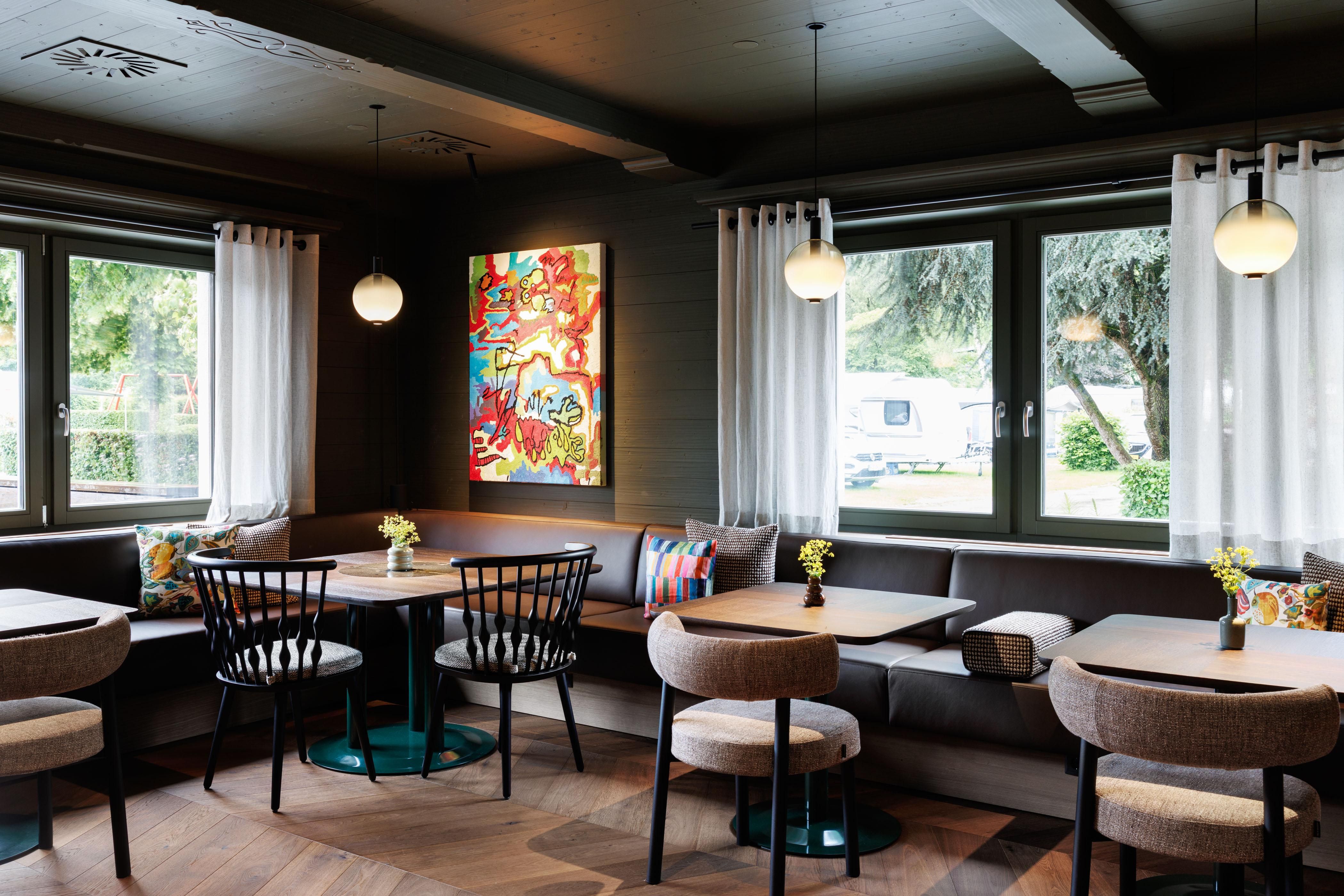 Ein modernes Café mit gemütlichen Sitzplätzen und bunten Dekorationen. An den Wänden hängen große Fenster, die viel Tageslicht hereinlassen.