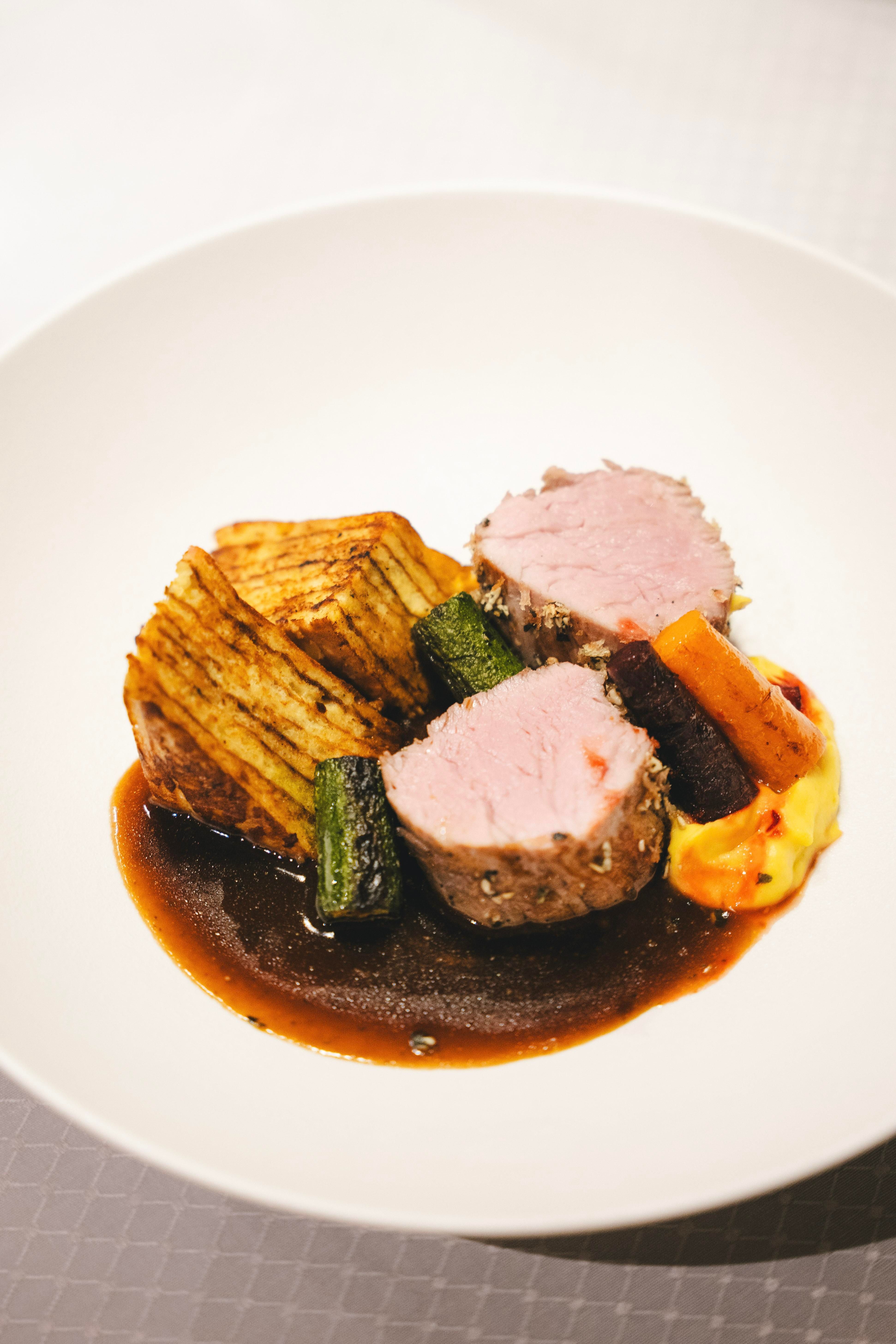 Ein kreativ angerichtetes Gericht mit zartem Fleisch, gebratenem Gemüse und einer dunklen Sauce. Dazu gibt es Kartoffeln in Form von Scheiben.