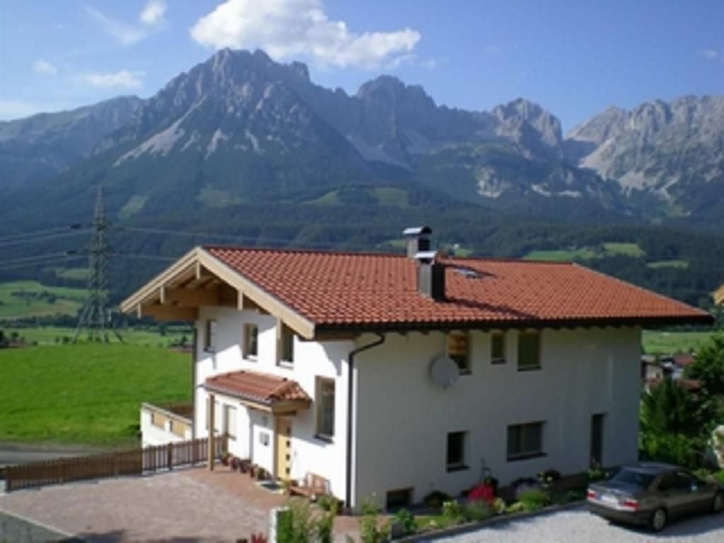 Ein schönes Haus mit rotem Dach in einer grünen Landschaft. Im Hintergrund erheben sich majestätische Berge unter einem blauen Himmel.