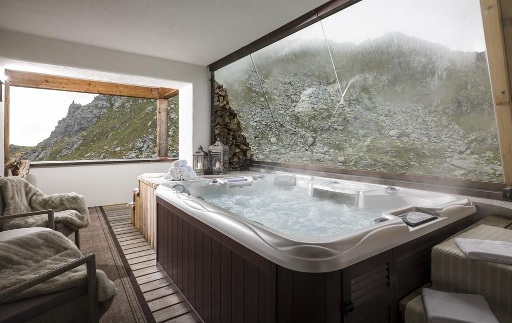 Ein Whirlpool in einem modernen Raum mit großen Fenstern bietet einen Blick auf eine felsige Berglandschaft. Der Raum ist komfortabel mit gemütlichen Möbeln und einem warmen Ambiente gestaltet.